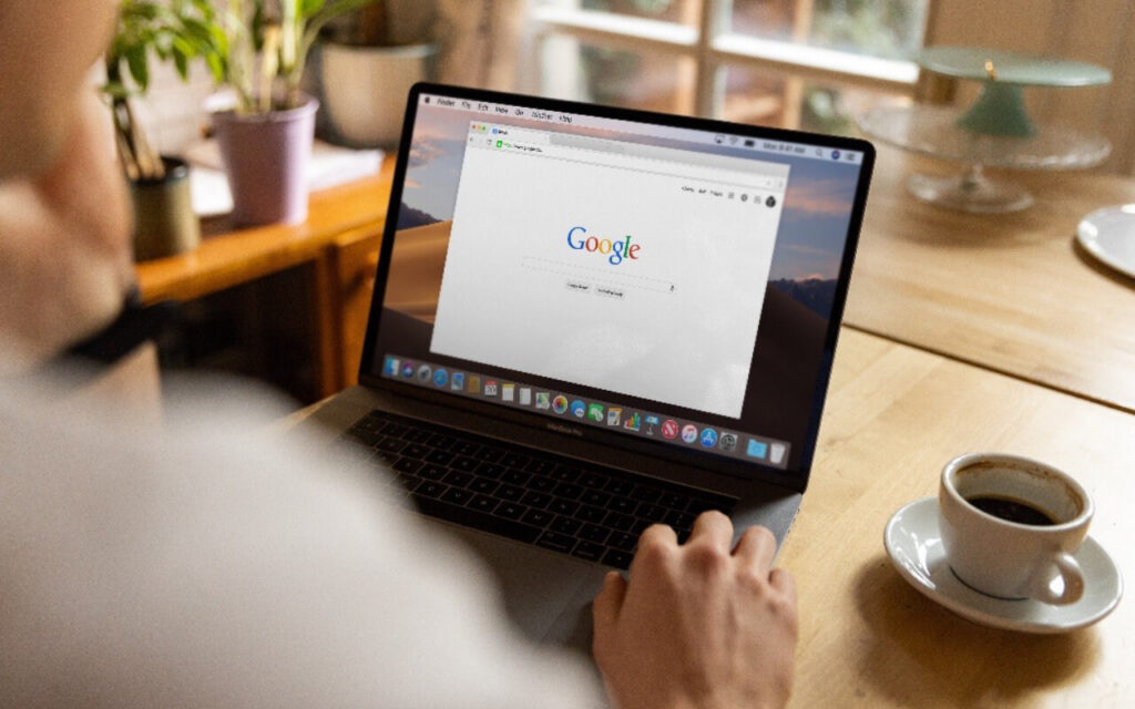 In einem gemütlichen Zimmer mit Topfpflanzen und einer Tasse Kaffee sitzt ein Mensch an einem Tisch mit seinem Laptop und hat Google geöffnet
